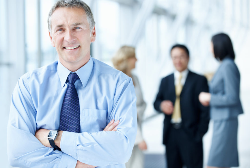 Portrait of a mature confident businessman standing with arms crossed and his colleagues in background